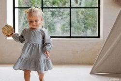 Petite fille blonde en robe grise jouant avec un objet, représentant les prénoms courts et jolis pour fille. Idéal pour les recherches sur les prénoms de bébé fille tendance et beaux. Liste de prénoms.